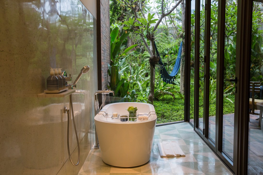 Modern bathroom with freestanding white bathtub overlooking lush garden vegetation through floor-to-ceiling windows.