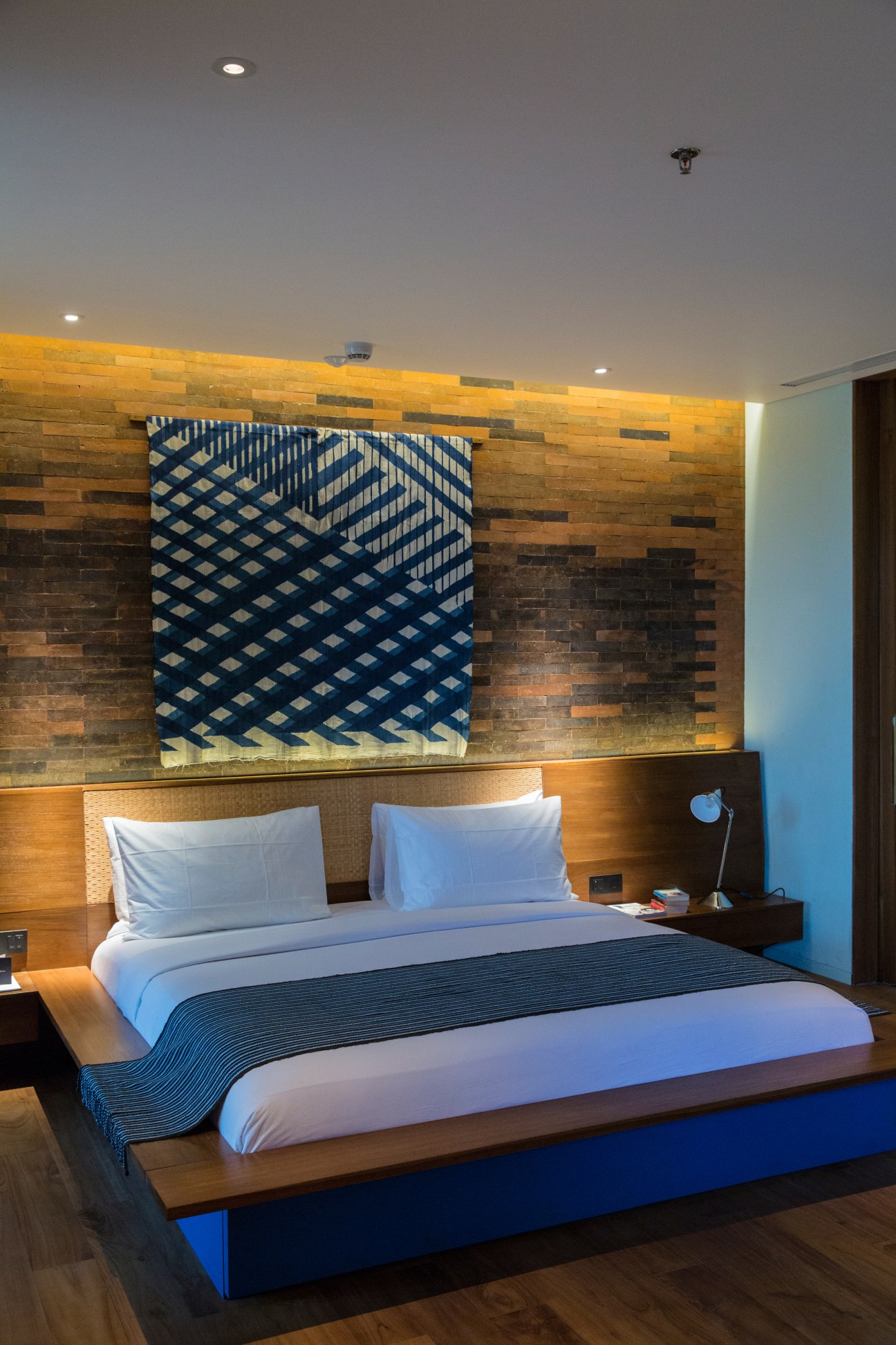 Modern bedroom with wooden platform bed, blue and white striped throw blanket, and indigo geometric tapestry on brick accent wall.