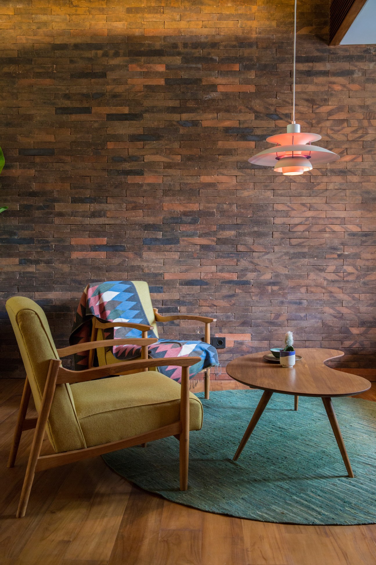 Mid-century modern lounge seating with wooden furniture, patterned textiles, and pendant lighting against a brick wall.