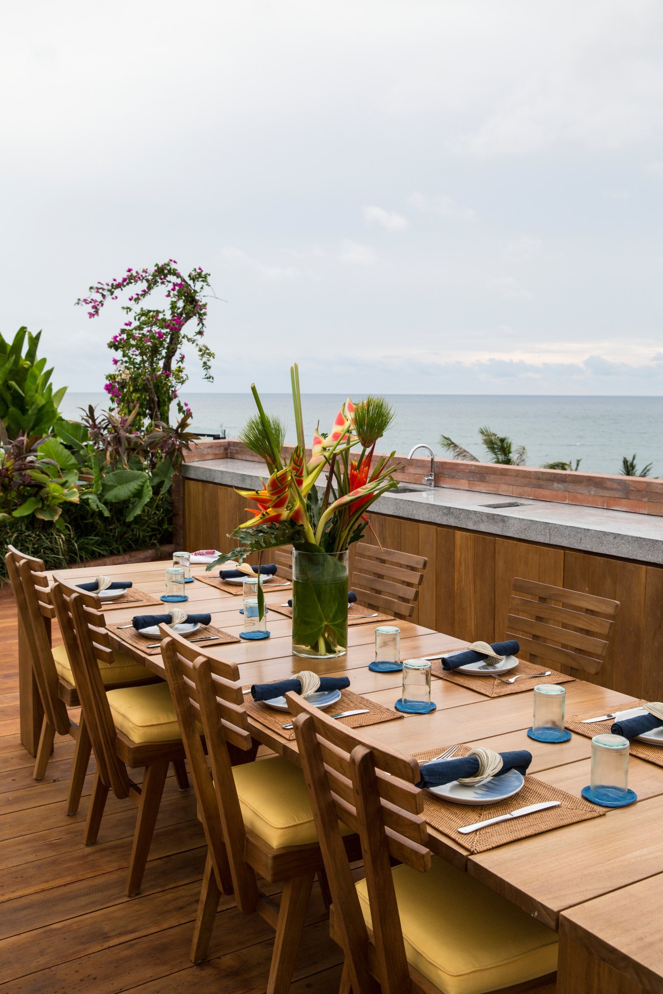 Wooden dining table set with place settings on a rooftop terrace overlooking the ocean, with tropical flowers and plants.