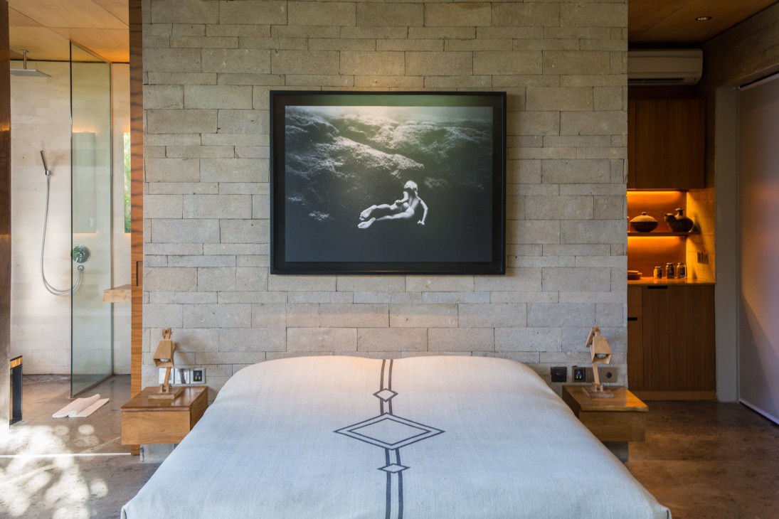 A minimalist hotel bedroom at The Slow with concrete block walls, white bedding, wooden nightstands, and framed artwork above the bed.
