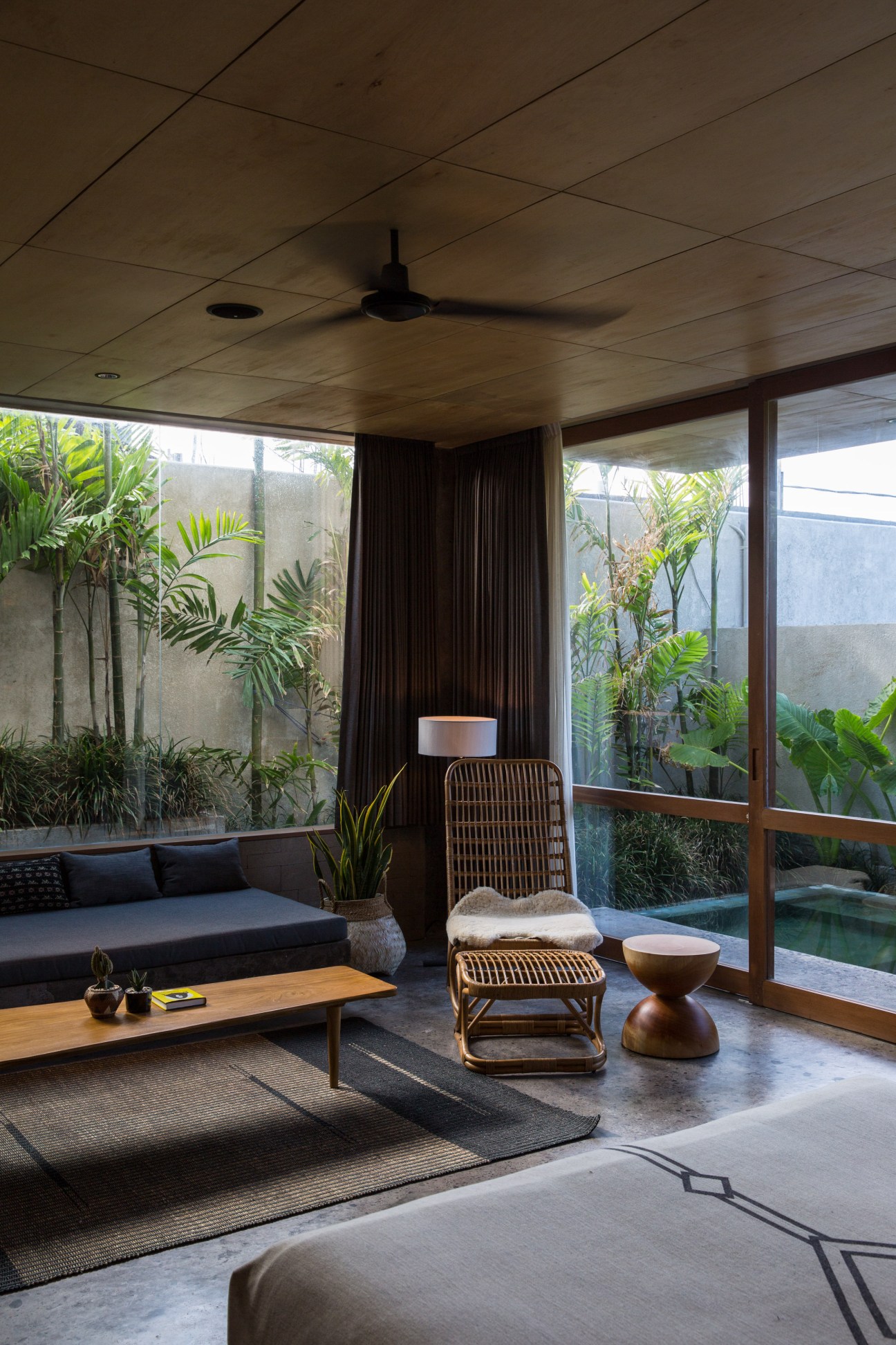Modern lounge room at The Slow hotel with rattan furniture, plants, and lush garden views through large windows.