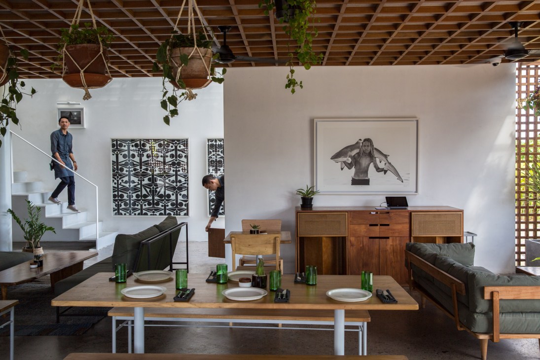 The Slow hotel's dining room features a long wooden communal table with place settings, mid-century furniture, artwork, and hanging plants.