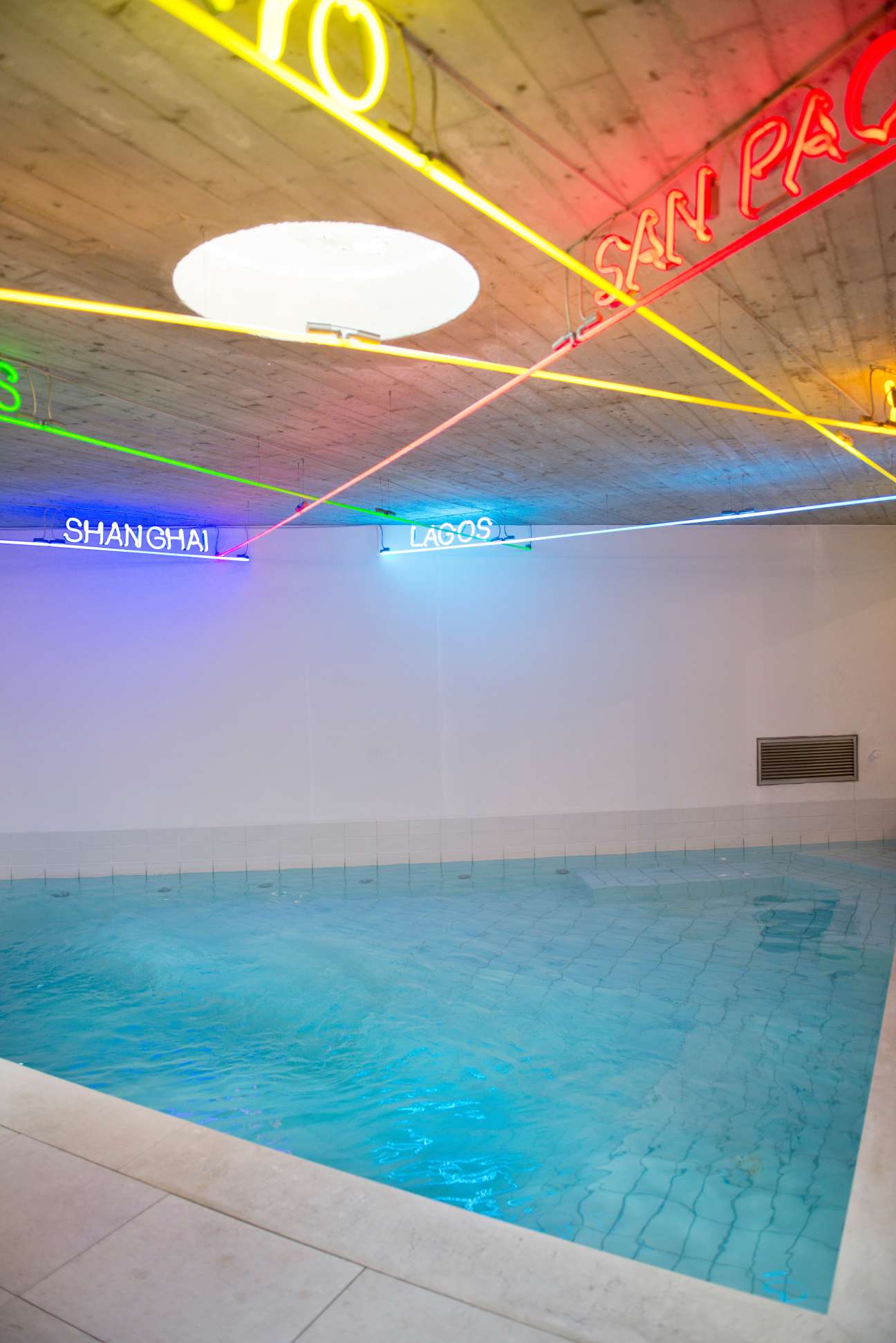 Neon light installation spelling city names above an indoor swimming pool