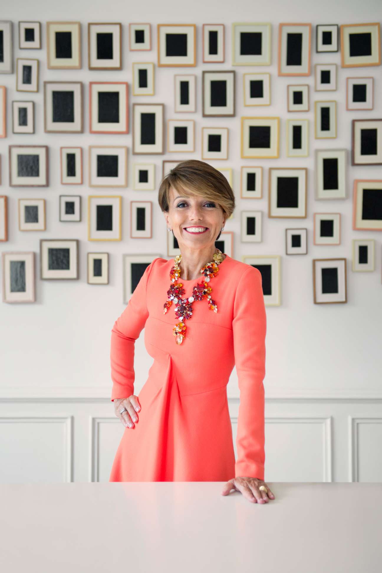 Woman in coral dress with statement necklace standing before wall of framed artworks