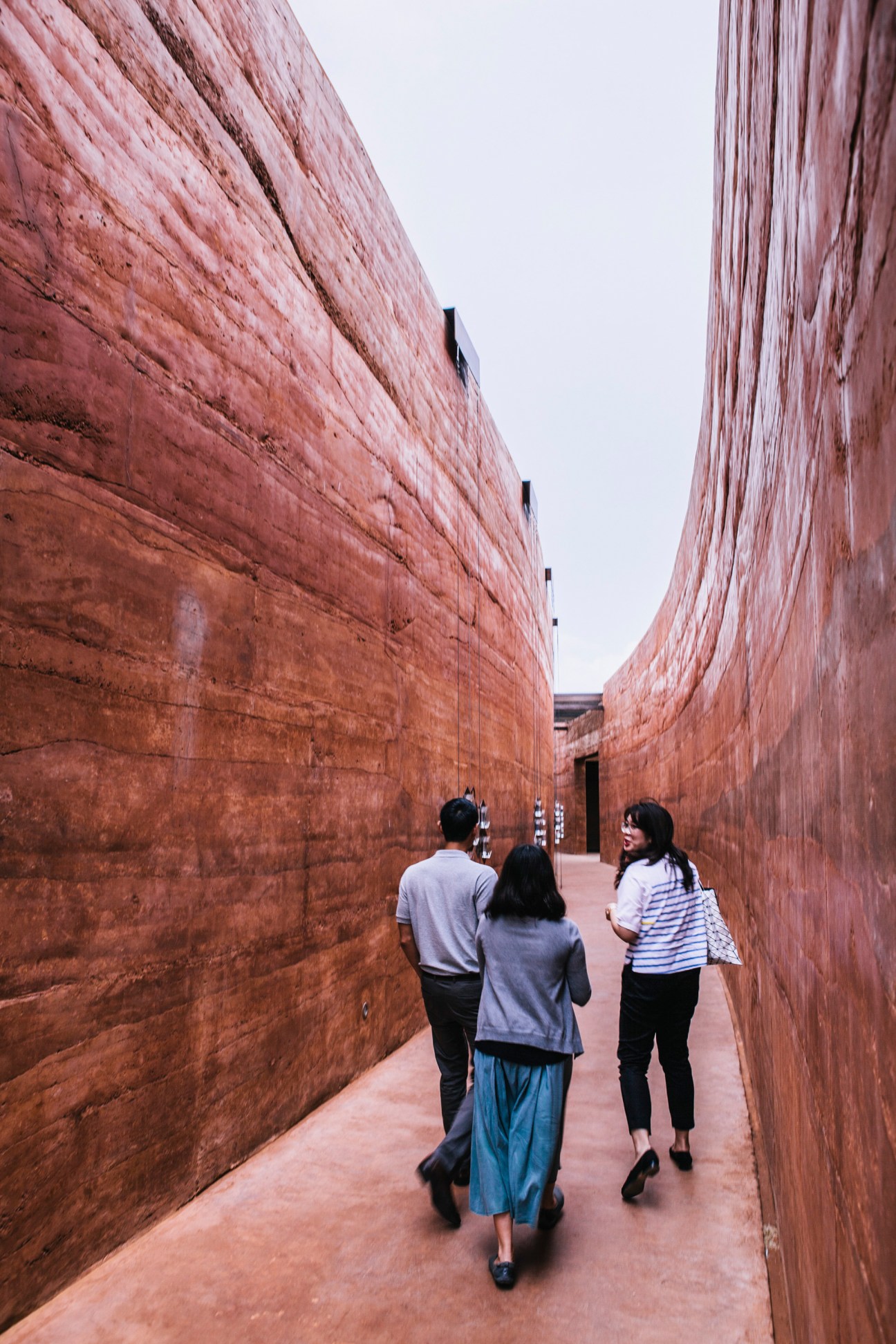 Three people walking through a narrow corridor with tall reddish-brown textured walls at PTT City Forest in Bangkok.