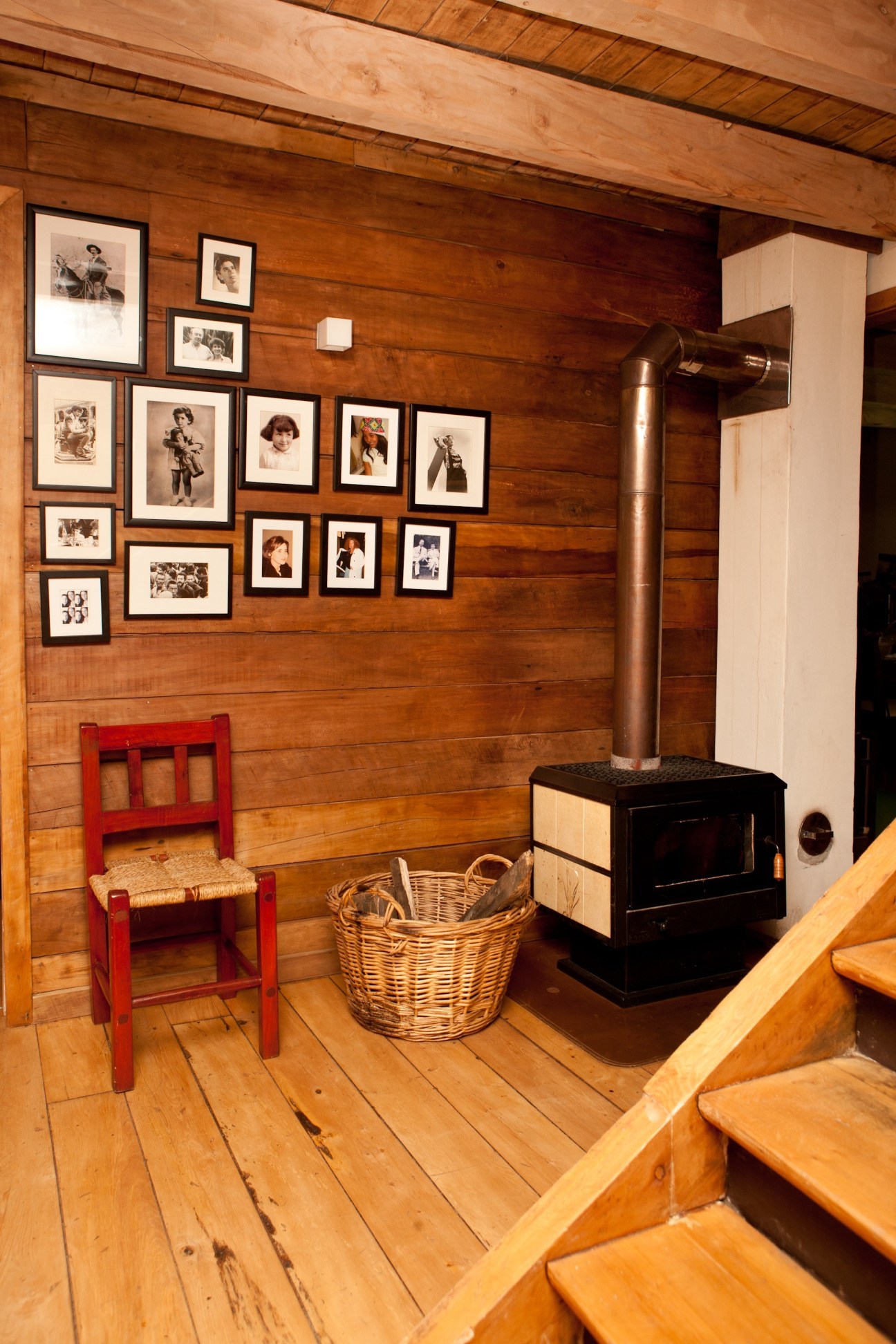 Interior of a wooden cabin with framed photographs on the wall, a red chair, wood stove, and wicker basket.