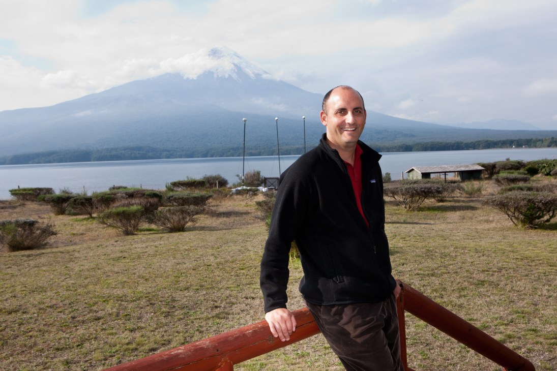 Enrique Papic standing by a wooden railing with snow-capped volcano and lake behind him.