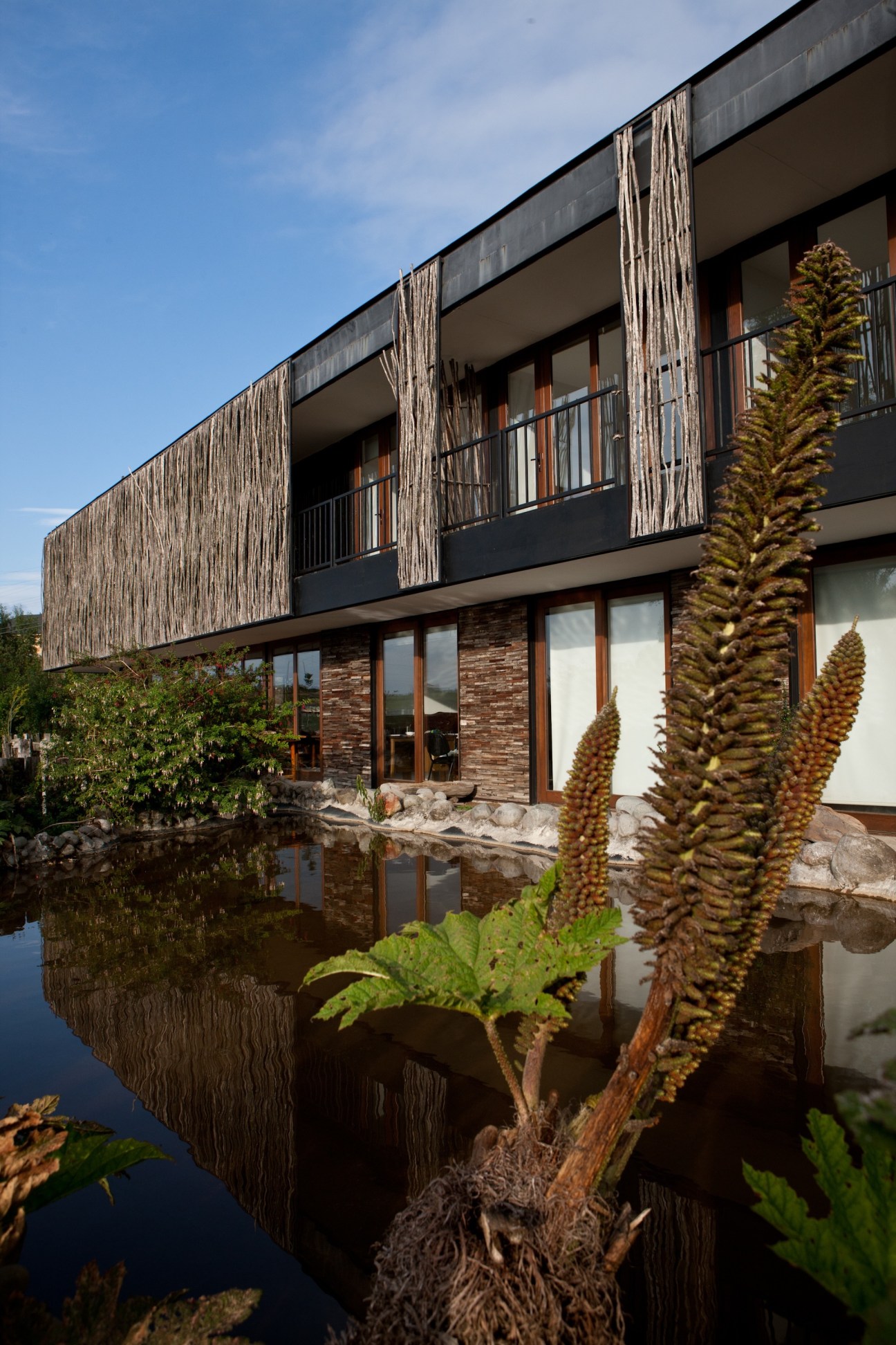 Modern hotel building with wood-sheathed facade and brick base reflected in still water, with native plants in foreground.
