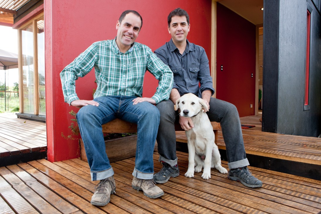 Two men sitting on a wooden deck with a white dog, against a red wall in a modern home interior.
