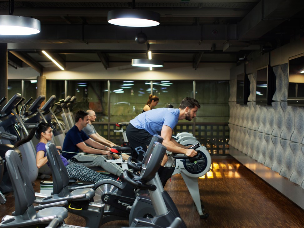 Multiple people exercising on stationary bikes and elliptical machines in a modern gym facility with bright overhead lighting.