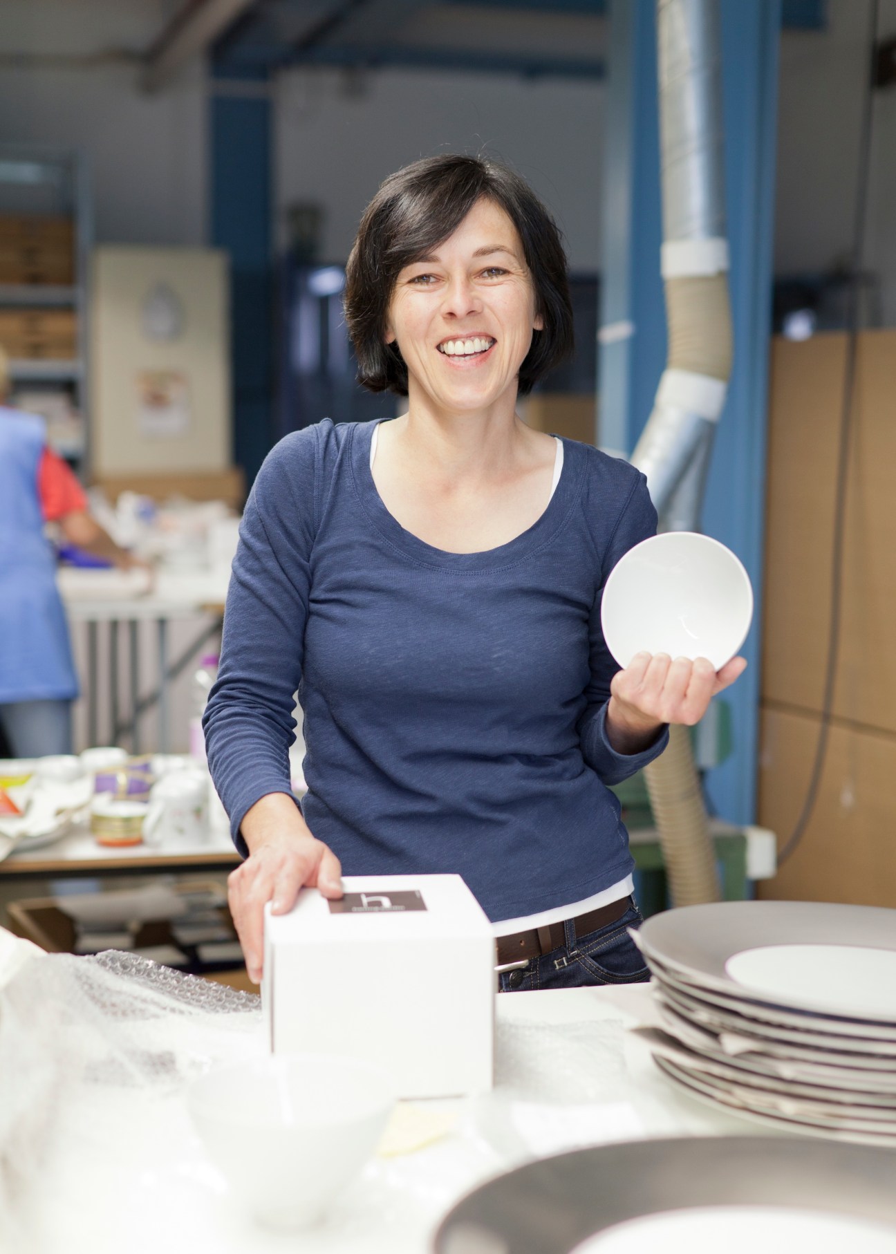 Woman holding a white porcelain plate in a factory workshop with stacked plates and packaging visible.