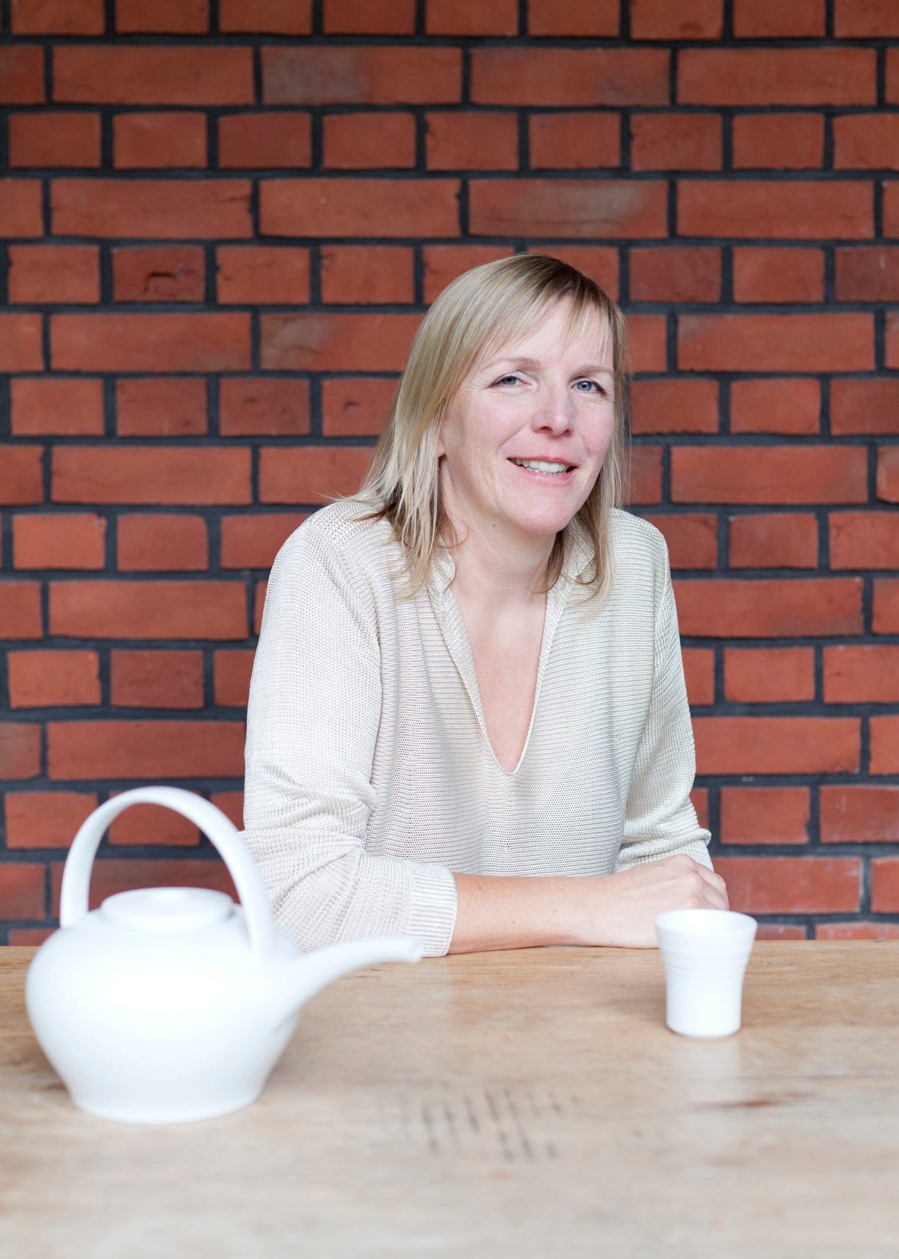 Stefanie Hering, founder of Hering Berlin porcelain, sits at a wooden table with white porcelain pieces against a red brick wall.