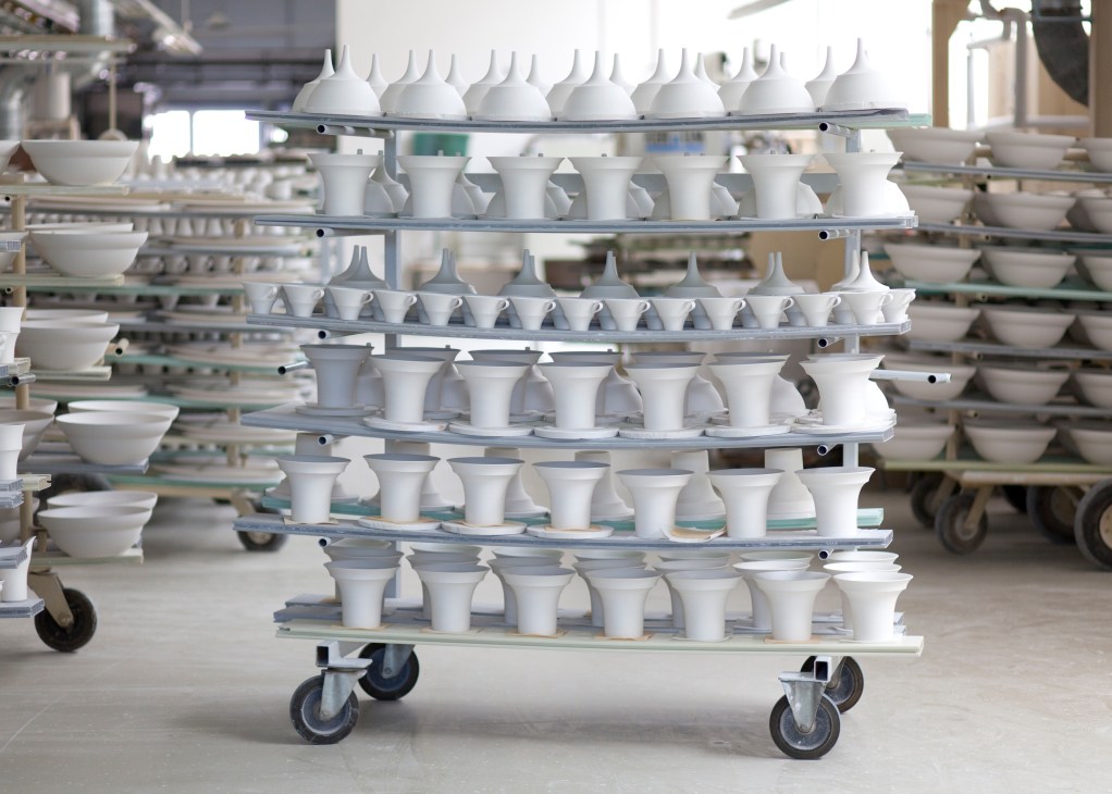 White porcelain bowls and cup forms arranged on metal shelving racks in a factory production area.