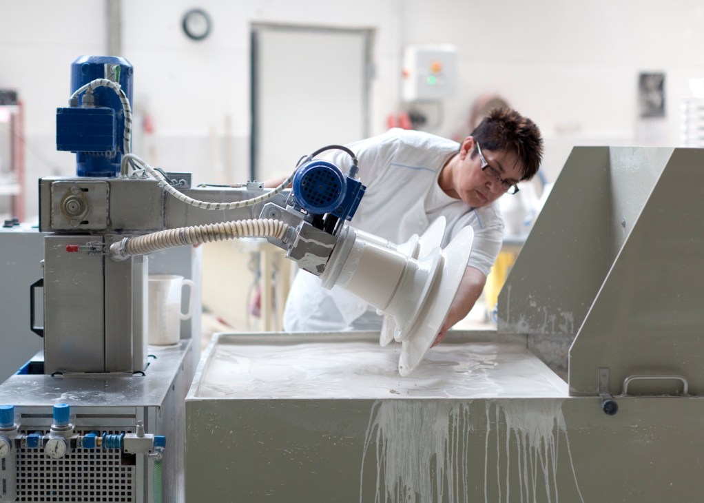 A worker in white shirt dips a white porcelain bowl into glaze using a machine with blue motor and hose.