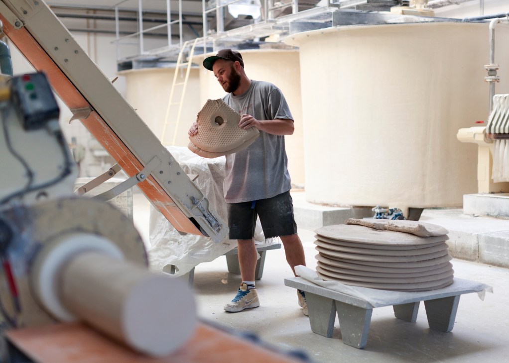Factory worker holding a perforated porcelain piece in production facility with stacked plates nearby.