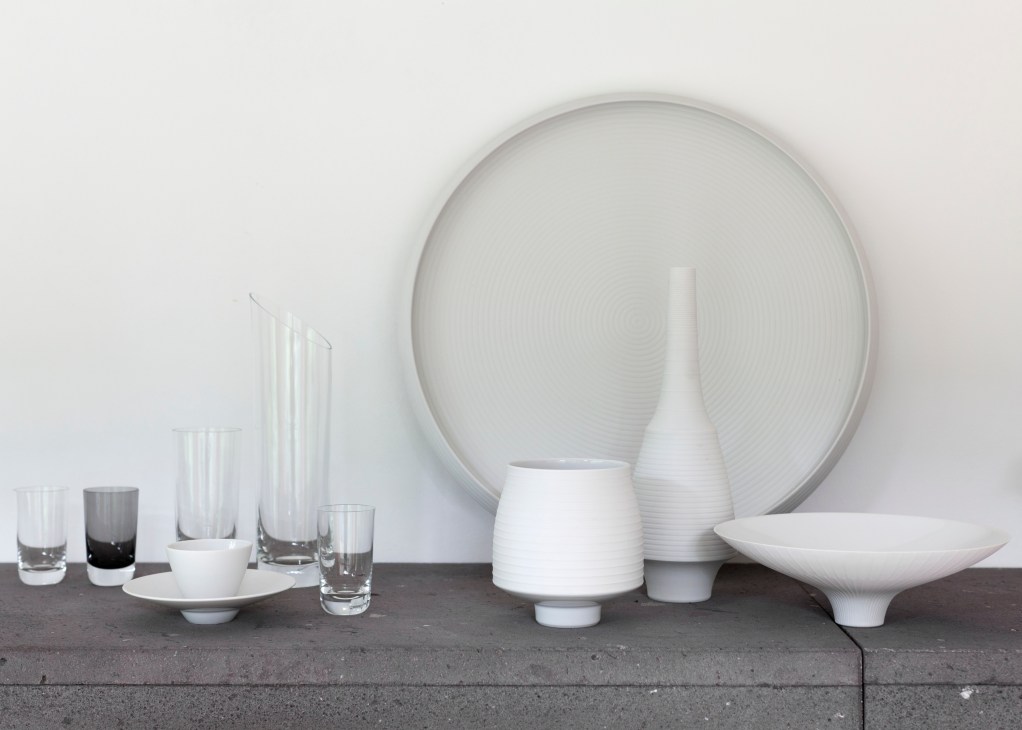 White porcelain tableware and glassware arranged on a dark surface against a white wall.