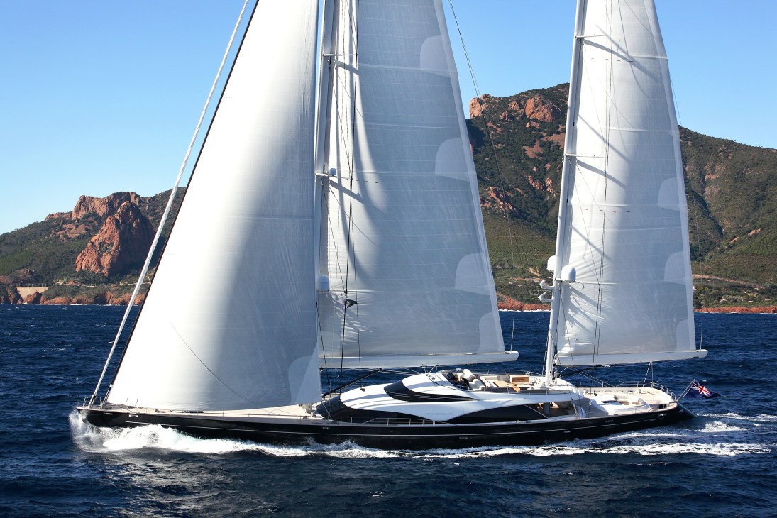 Luxury sailing yacht Twizzle with white sails deployed on blue water near rocky Mediterranean coastline.