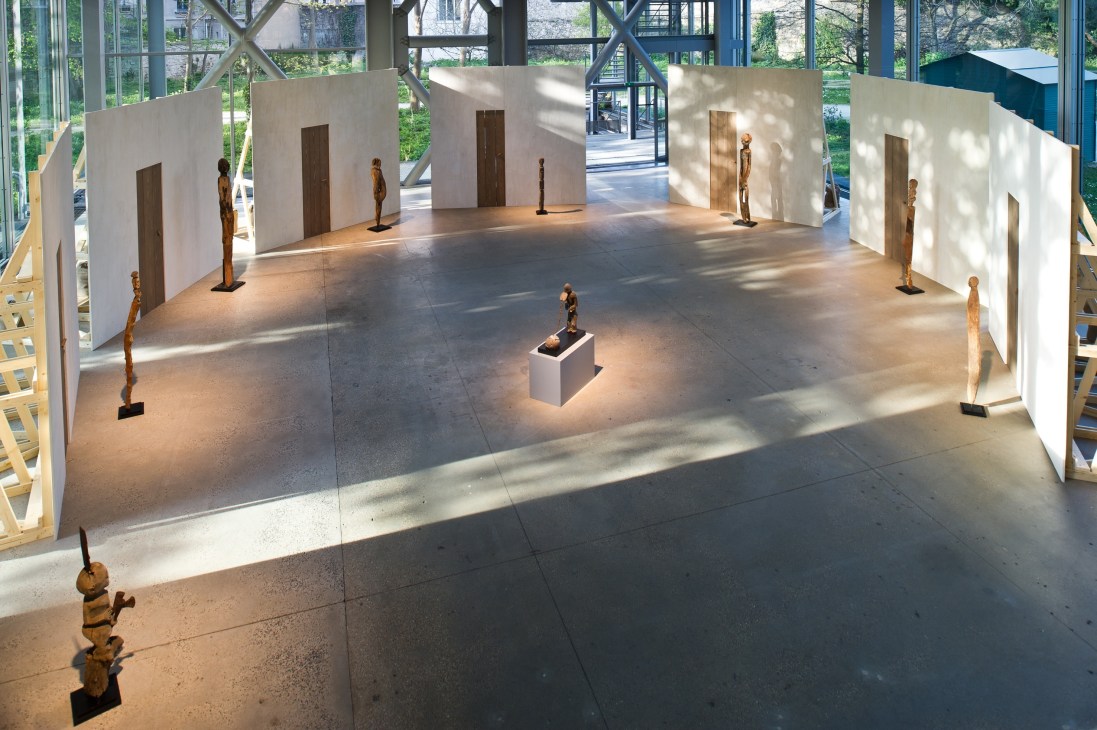 Art gallery interior with white walls displaying wooden sculptures on pedestals in natural light.