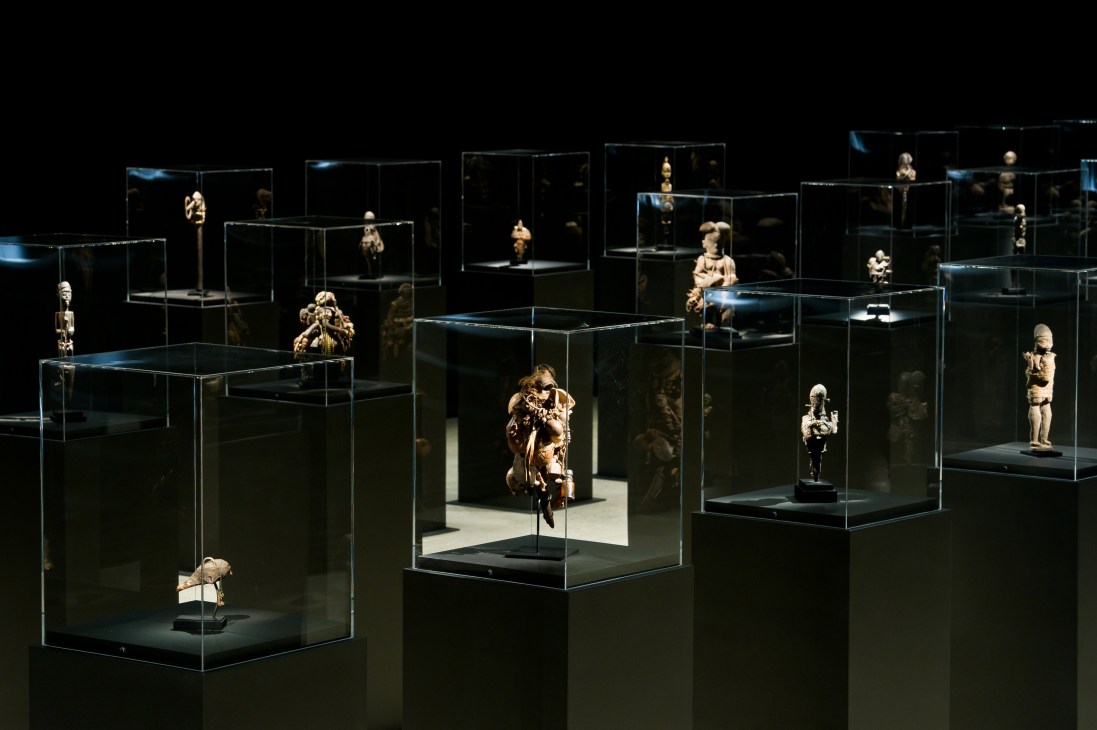 Museum display of West African voodoo art sculptures in glass cases with dramatic lighting against dark background.