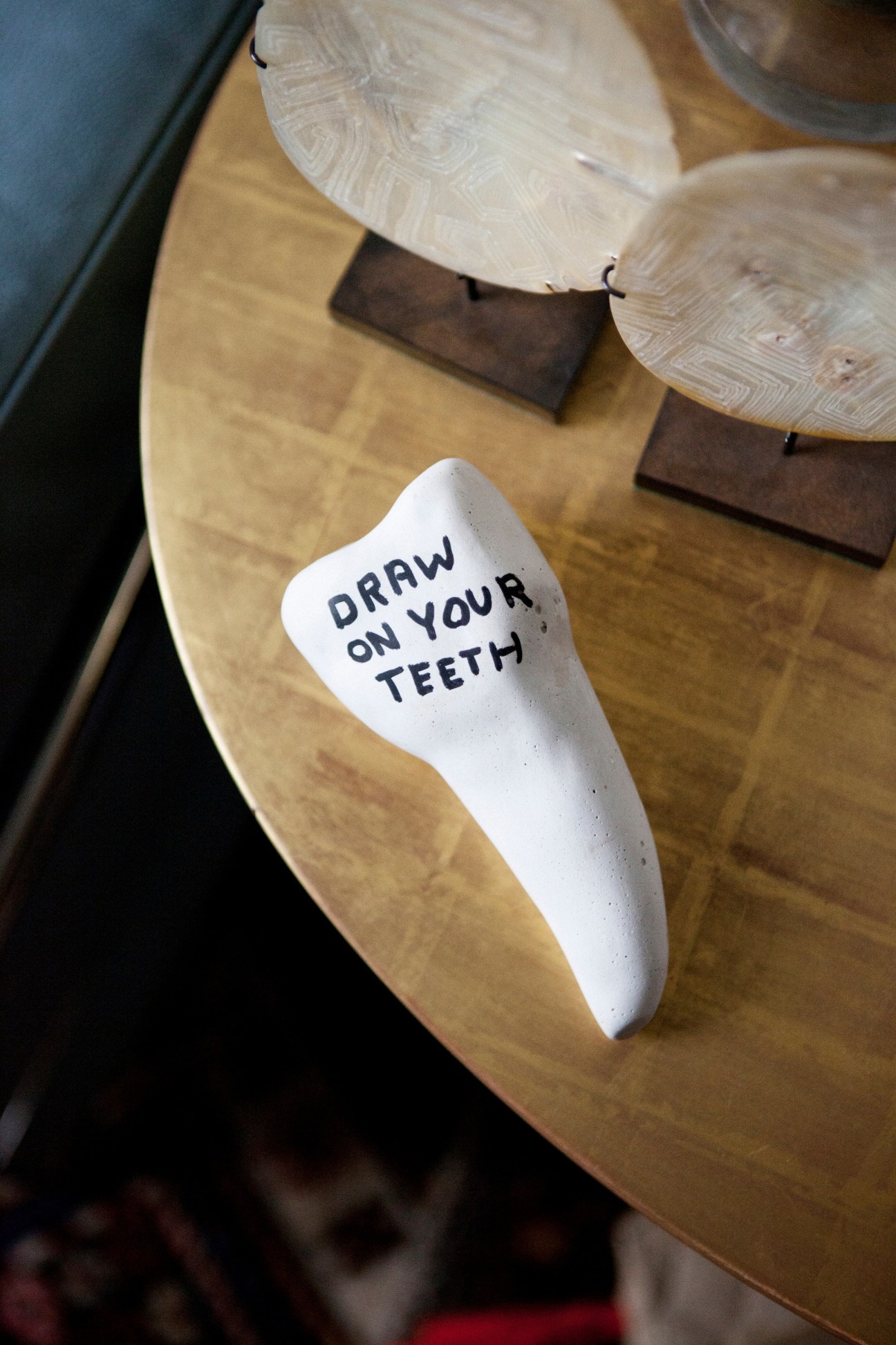 White ceramic tooth sculpture with "DRAW ON YOUR TEETH" text on a wooden table with decorative shells.