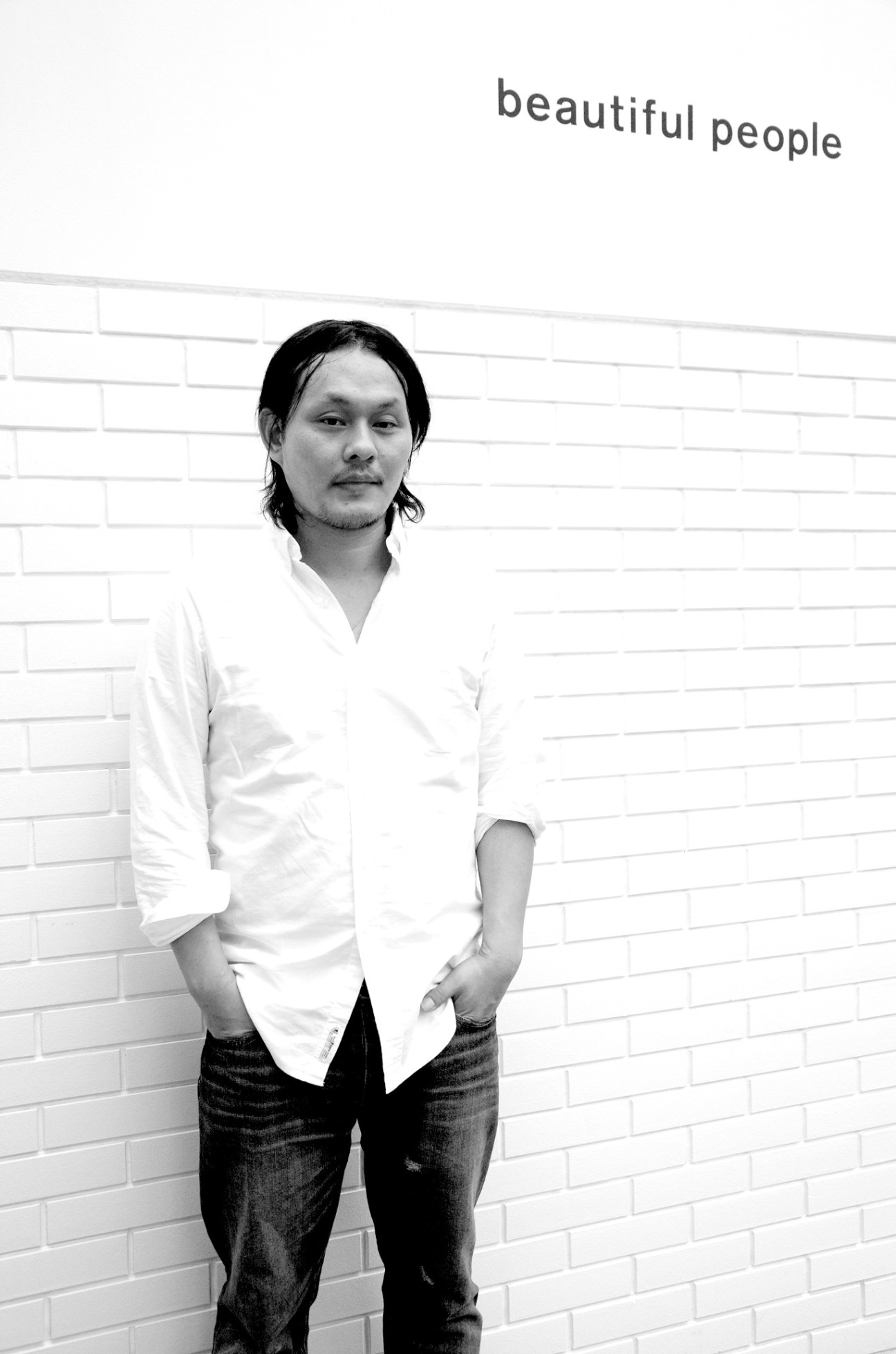 Hideriori Kumarkiri, founder of Beautiful People, stands against a white brick wall wearing a white shirt and dark jeans.