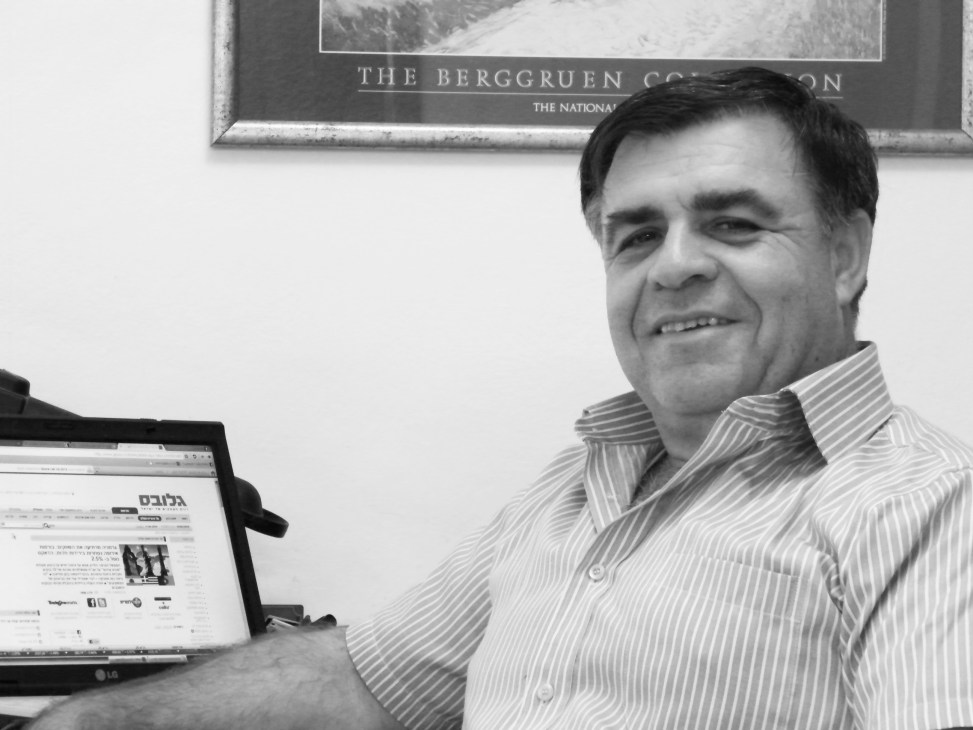 Urban planner Zvika Mintz smiling at his desk with a laptop in his office, with framed artwork on the wall behind him.