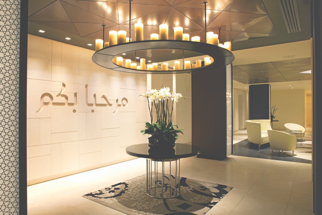 Interior of Qatar Airways lounge with Arabic calligraphy on wall, circular candle chandelier, and cream-colored seating area.