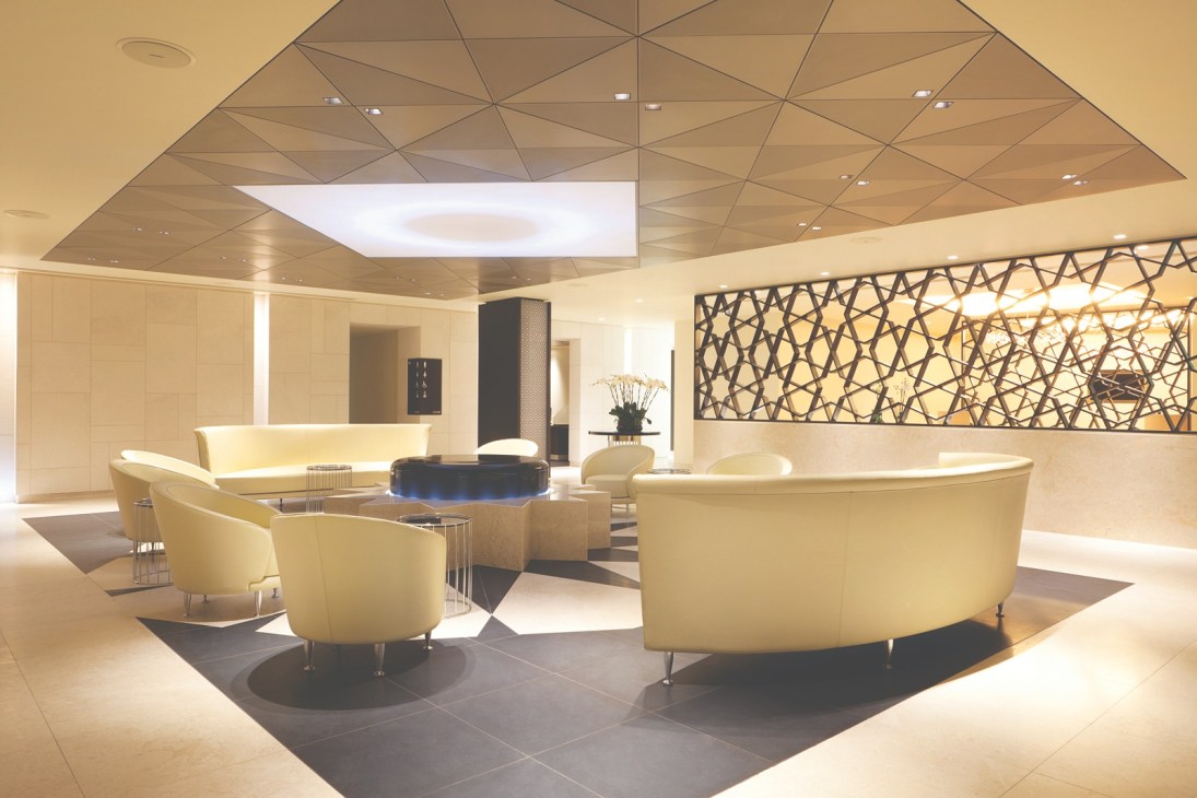 Modern airport lounge interior with cream-colored curved seating, geometric ceiling panels, and Islamic-inspired fretwork wall decoration.