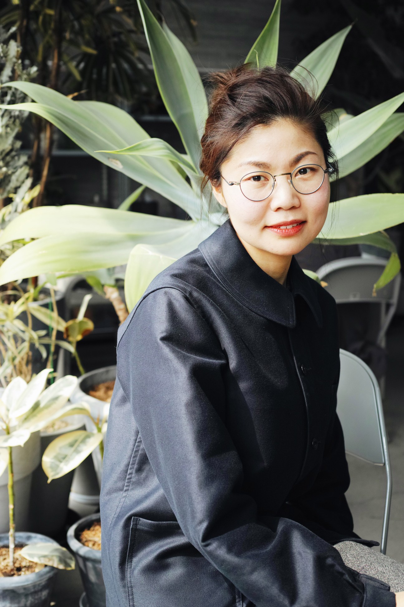 Kwon Kyehee, planning department manager, poses among potted plants in a dark blazer and round glasses.