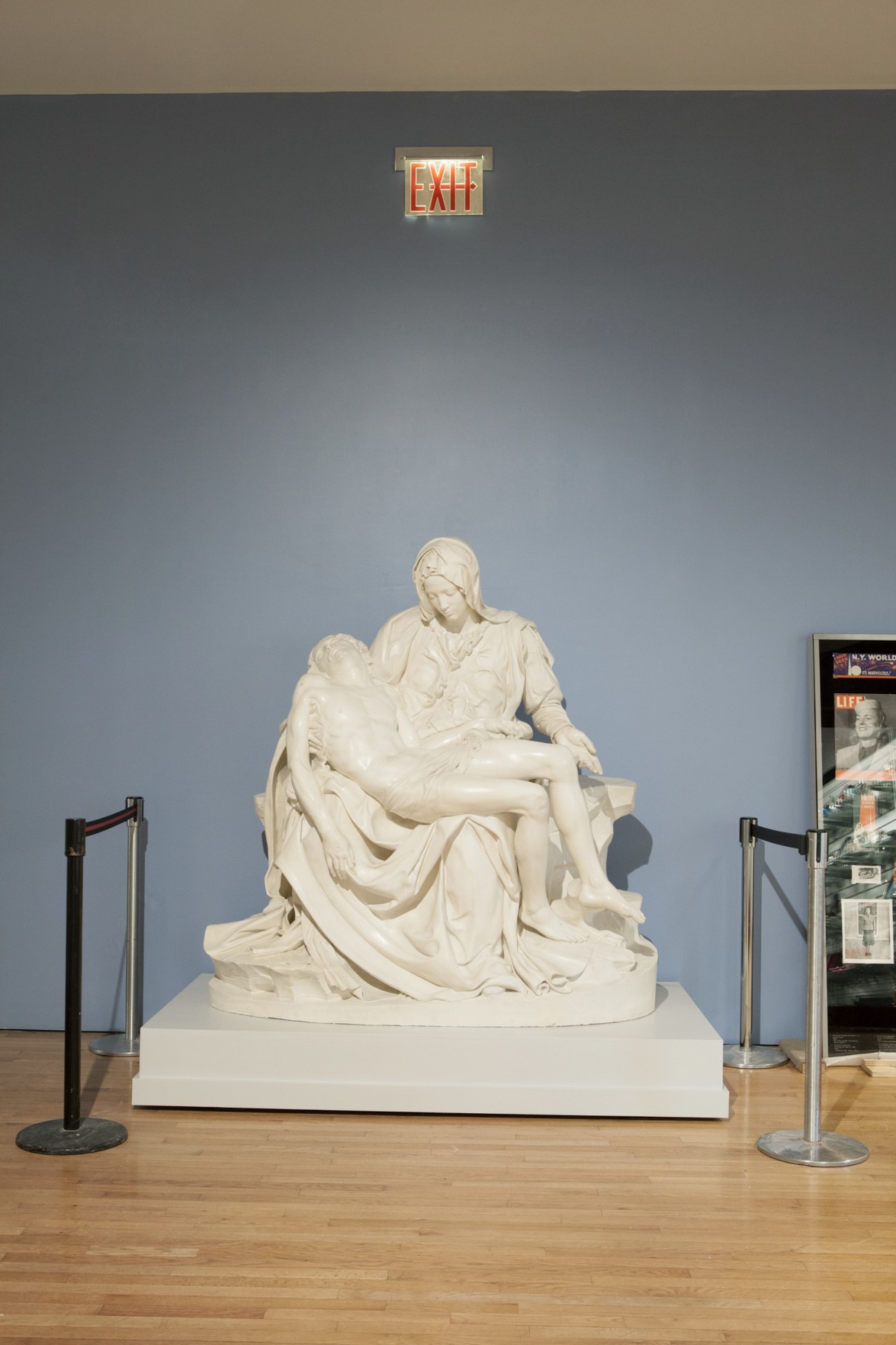 Plaster cast of Michelangelo's Pieta displayed on a white pedestal in a gallery with blue walls and an exit sign above.