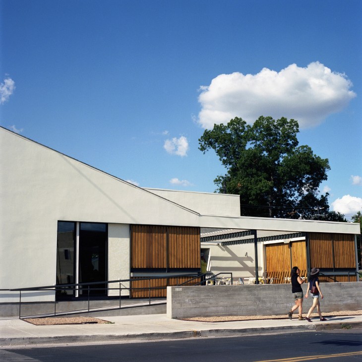 Modern white building with wooden slats and flat roof, two pedestrians walking past on street.