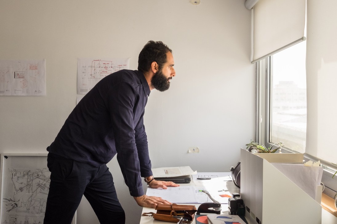 Tarik Al Zaharna in his studio, leaning over architectural drawings at a desk with sketches pinned on the wall behind him.