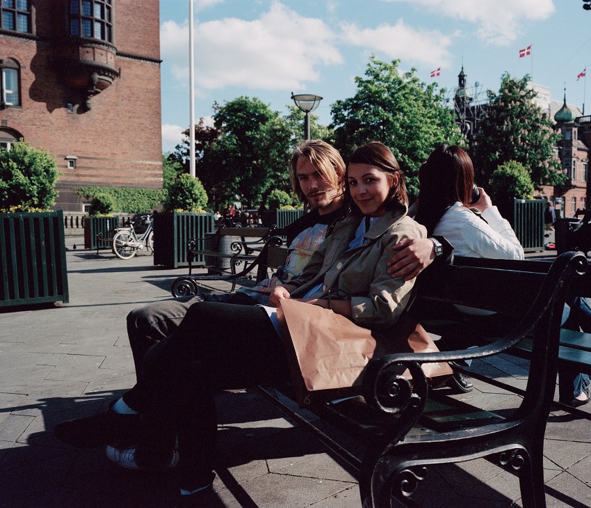 A couple sitting on a bench in Rådhuspladsen square in Copenhagen, with historic architecture and cyclists visible in the background.