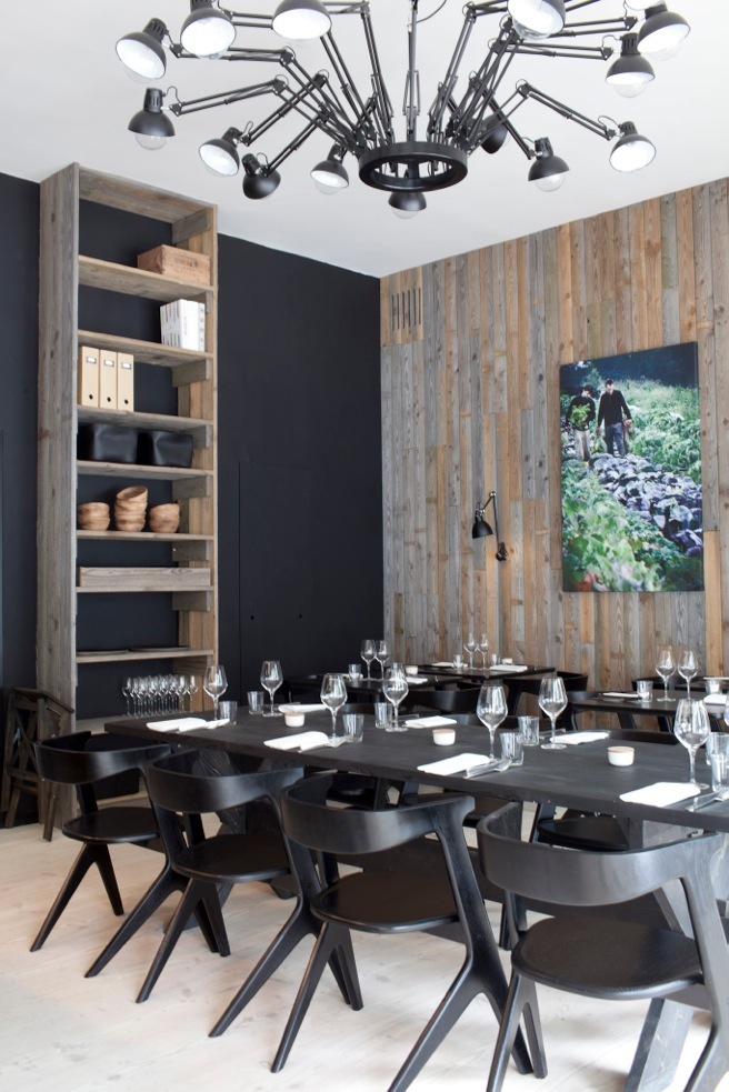 Modern restaurant dining room with black table, chairs, wooden wall paneling and industrial ceiling lights.