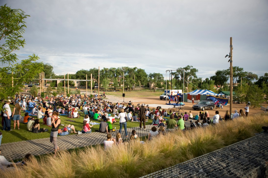 Crowds of people gather on grass in Railyard's public park with native plantings and wooden structures.