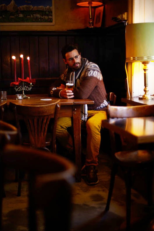 Man seated at a bar table wearing designer clothing including a Jejia jumper, Visvim shirt, and Bottega Veneta trousers.
