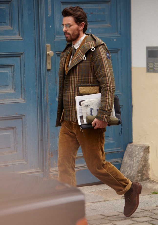 Model wearing plaid coat, Comme des Garçons jacket and trousers, carrying laptop case outside blue door.