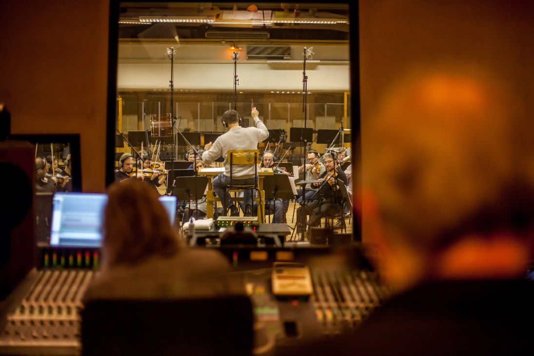 Conductor leading string musicians during a recording session in a studio, viewed through a control room window with equipment in foreground.