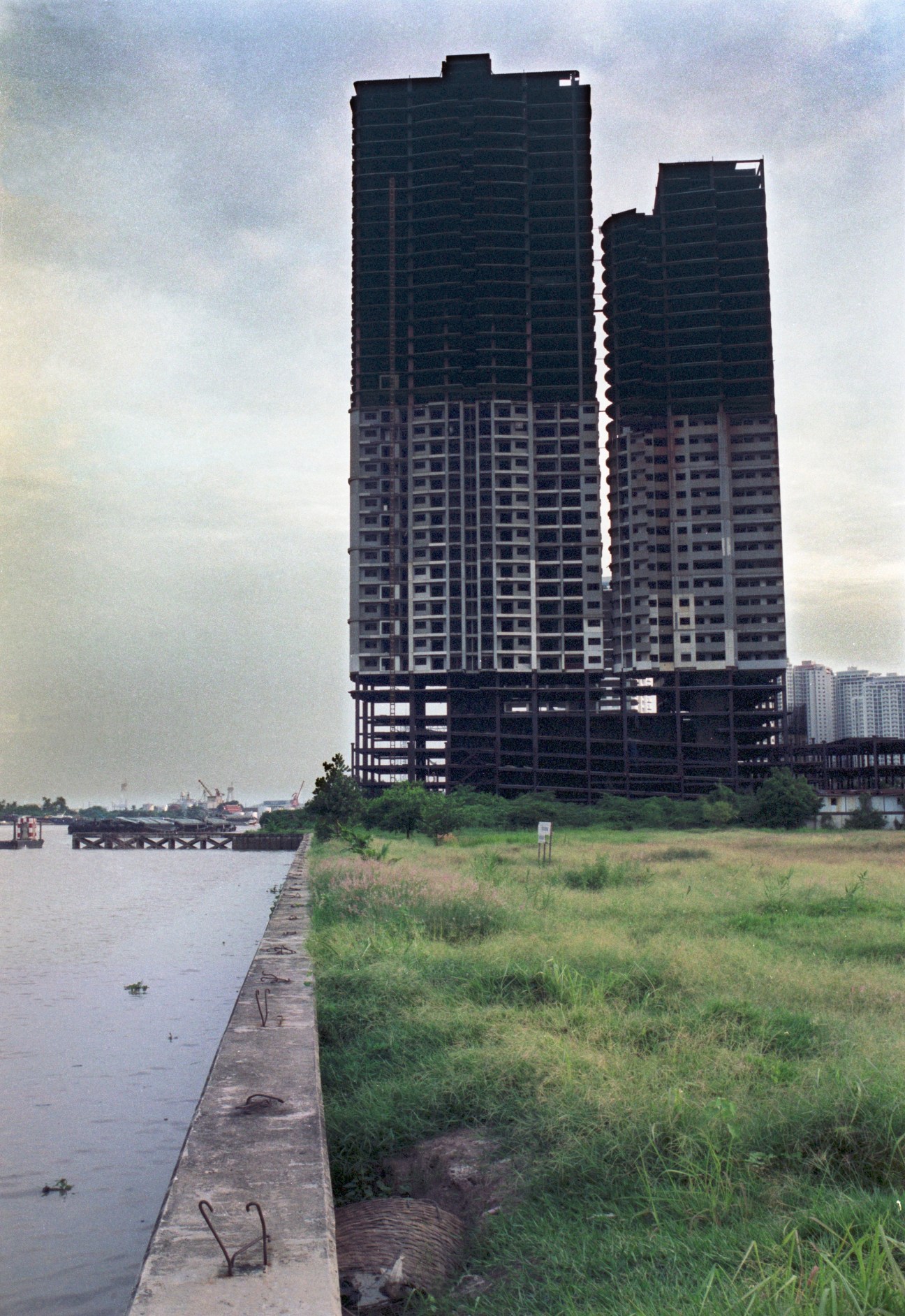 Unfinished high-rise towers in Bangkok with exposed concrete structure and glass facade alongside water and overgrown grass.
