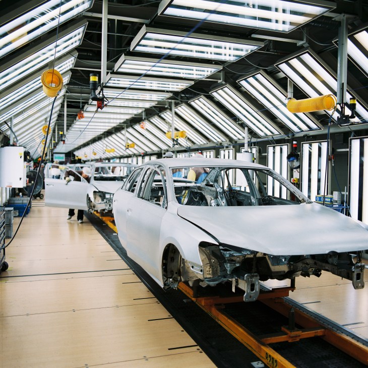 Volkswagen car assembly line with white vehicles under bright skylights in manufacturing facility.