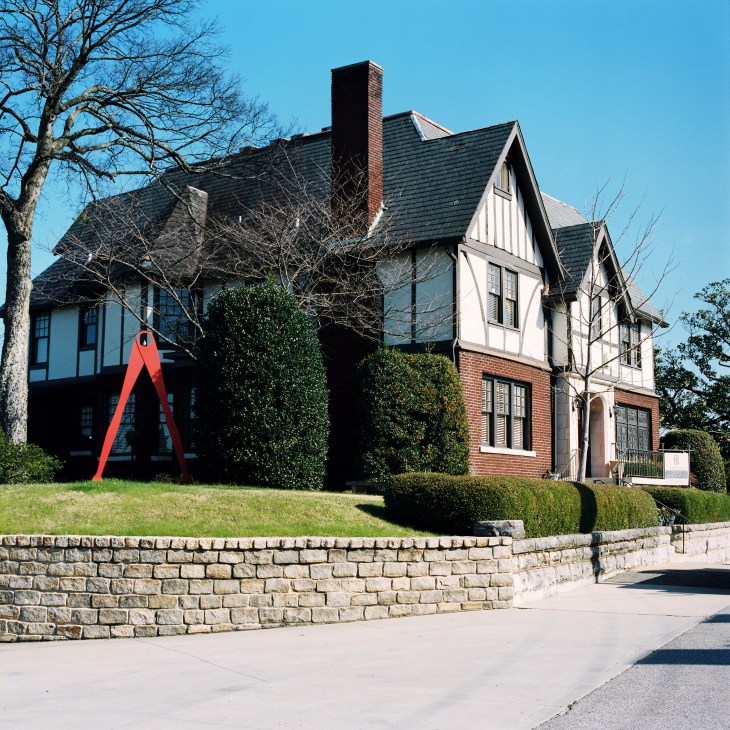 Historic house with red modern art sculpture in front, surrounded by manicured landscaping and stone wall.