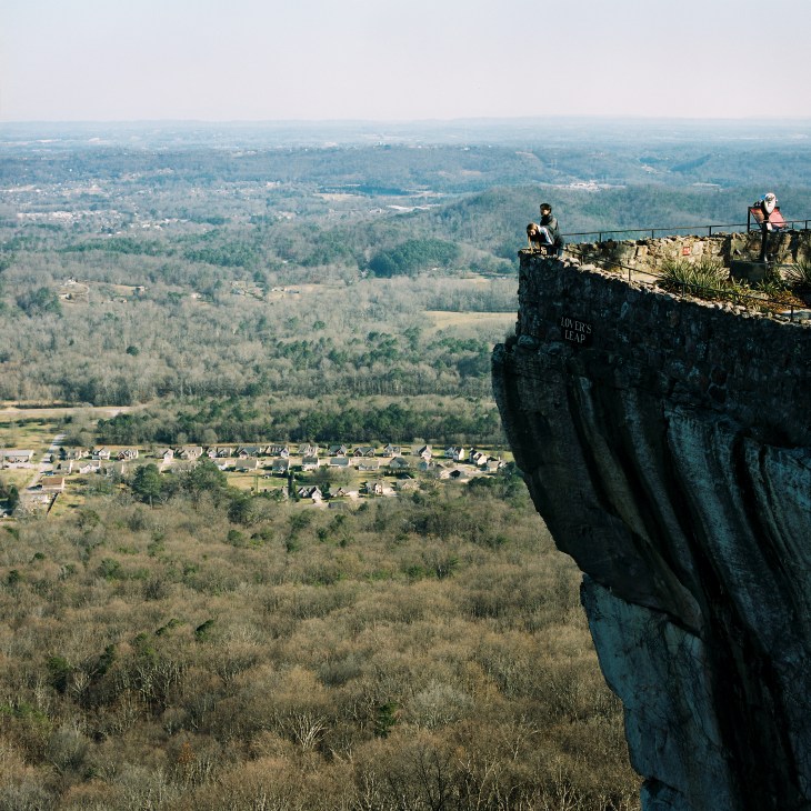 Hikers stand atop a rocky cliff at Lover's Leap on Lookout Mountain overlooking forested valleys and a residential area below.