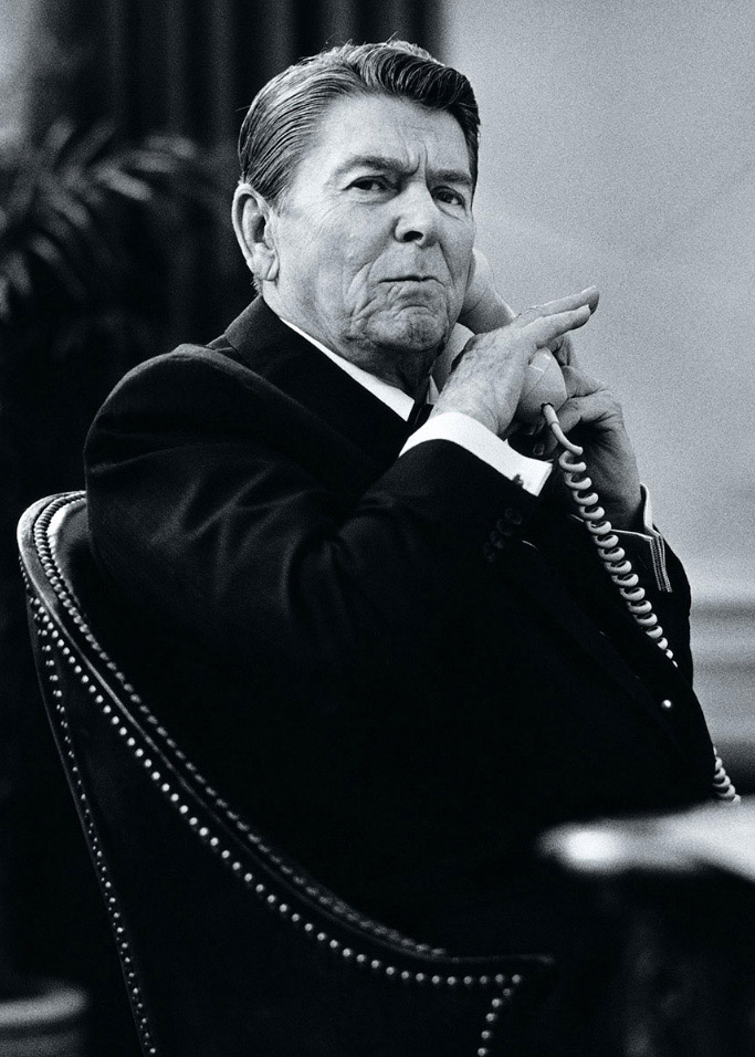 Ronald Reagan sitting in chair with hand to chin, speaking on telephone in black and white photograph.