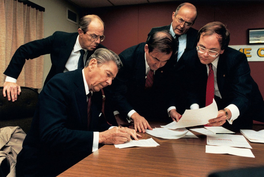 Ronald Reagan signing documents at a table surrounded by five men in suits in an office setting.