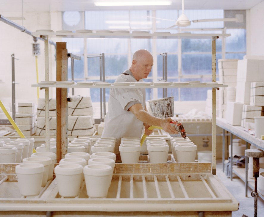 Worker applying decorative detail to white ceramic vessels in a bright pottery studio.