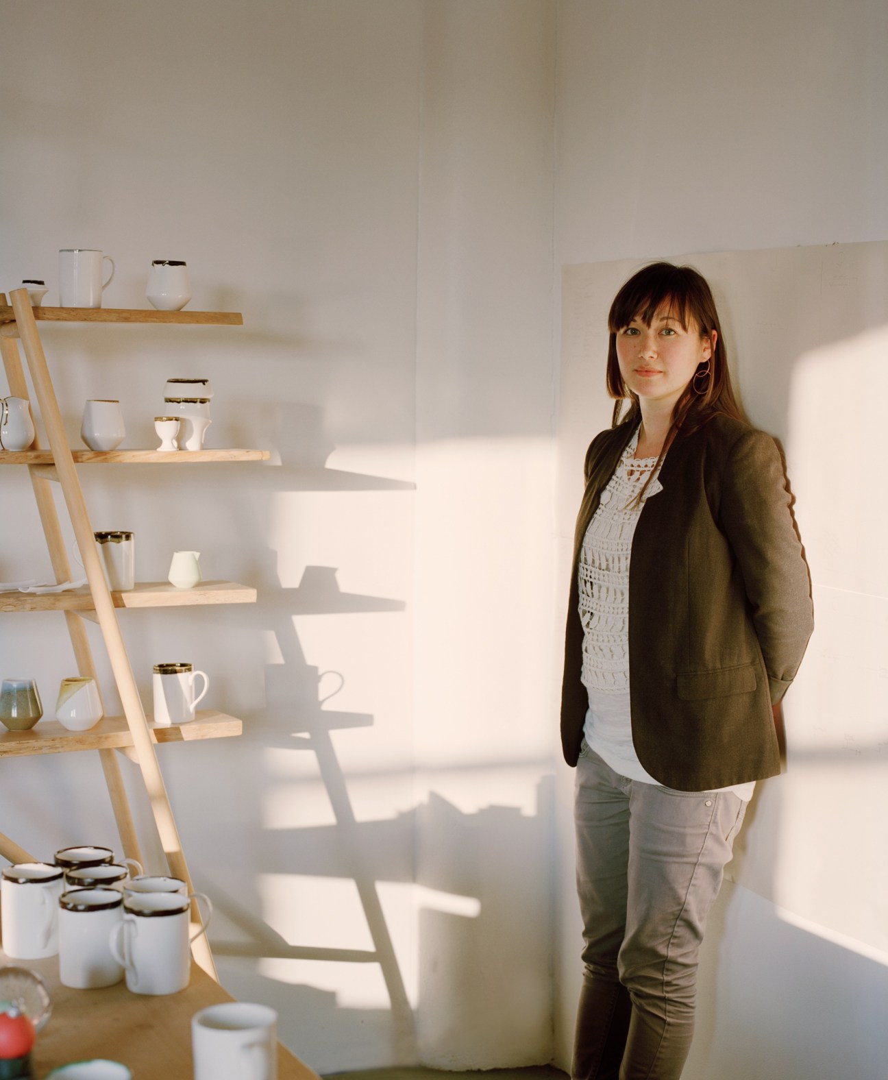 Reiko Kaneko stands in her bright studio with ceramic vessels displayed on wooden shelves behind her.