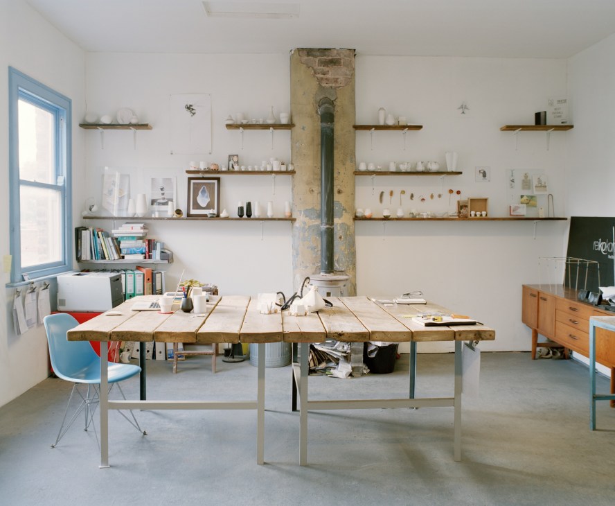 Ceramicist's studio workspace with wooden work table, shelving displaying ceramic pieces, and large industrial chimney structure.