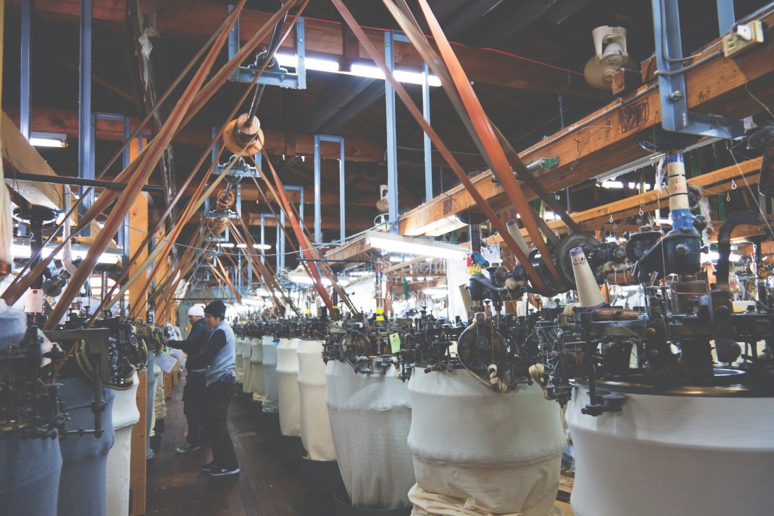 Factory workers monitor loopwheel circular knitting machines producing white cotton fabric in an industrial facility.