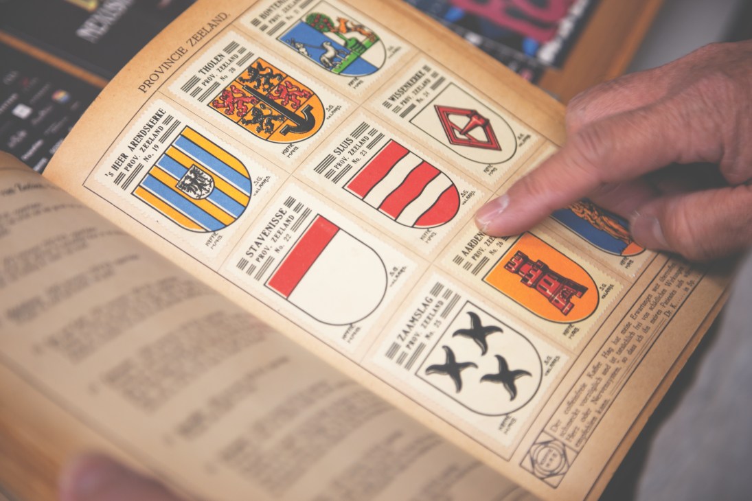 A hand flips through a vintage book displaying colorful Dutch heraldic crests and coat of arms designs.